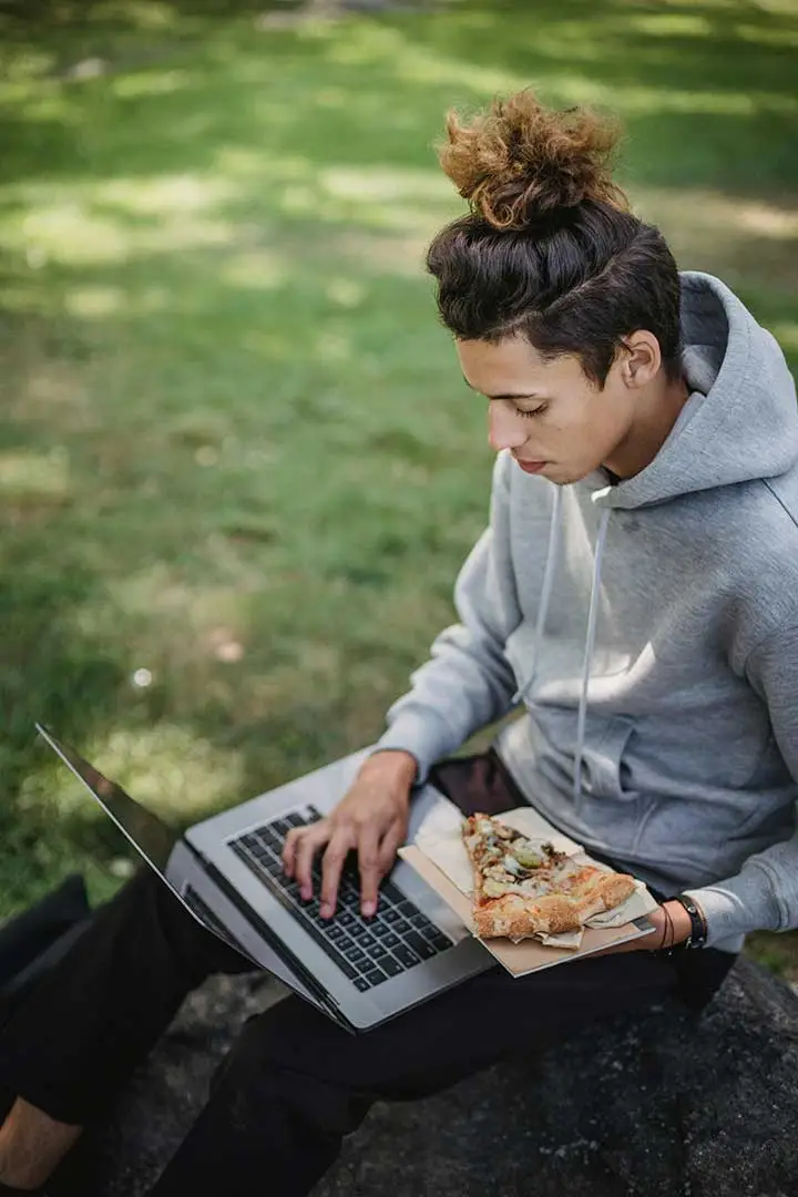 Student on laptop while holding a piece of pizza