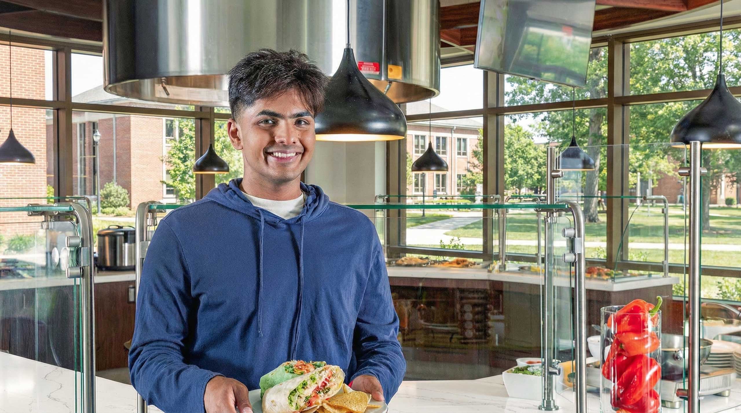 Male student smiling and holding a plate of food with serving line and large windows in the background